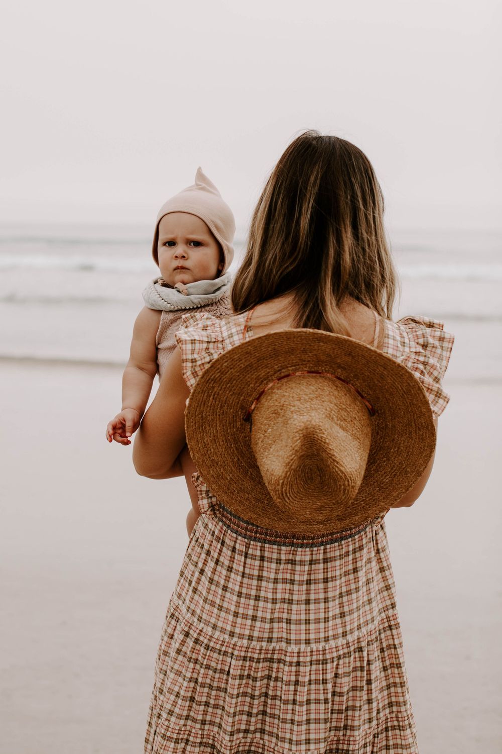 Person in plaid dress carrying baby while walking along sandy beach wearing brown sunhat.