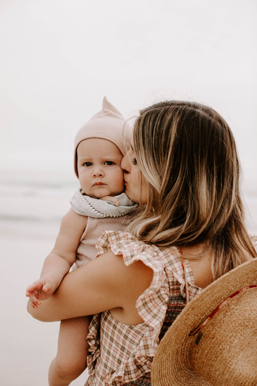 Parent in plaid dress holds and embraces young child against bright beach backdrop.