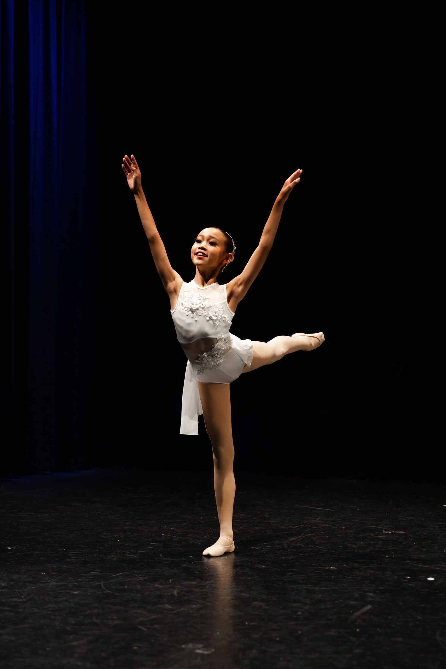 Ballet dancer in white tutu and pointe shoes performs graceful movements on dark stage during solo performance.