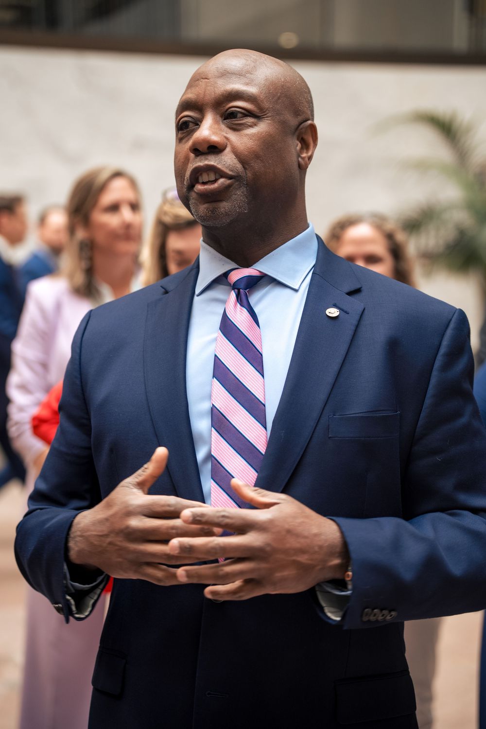A man in a navy suit with a striped tie using expressive hand gestures while speaking.