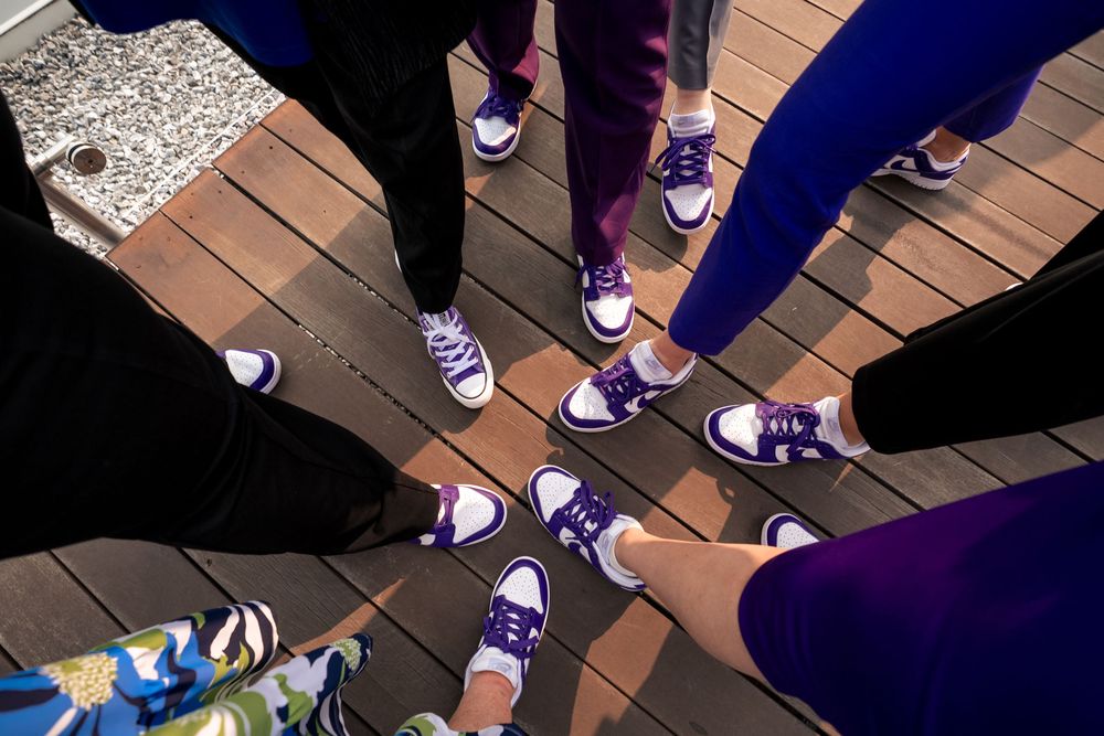A circle of people wearing matching purple Converse sneakers standing on wooden decking.