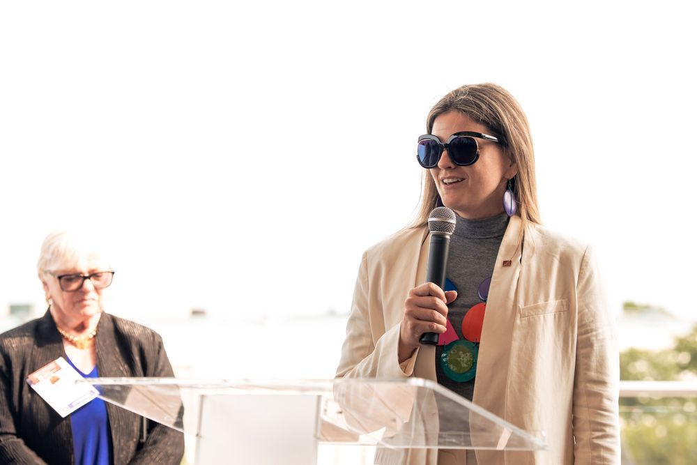 A woman in a beige coat and sunglasses speaking at a podium outdoors.