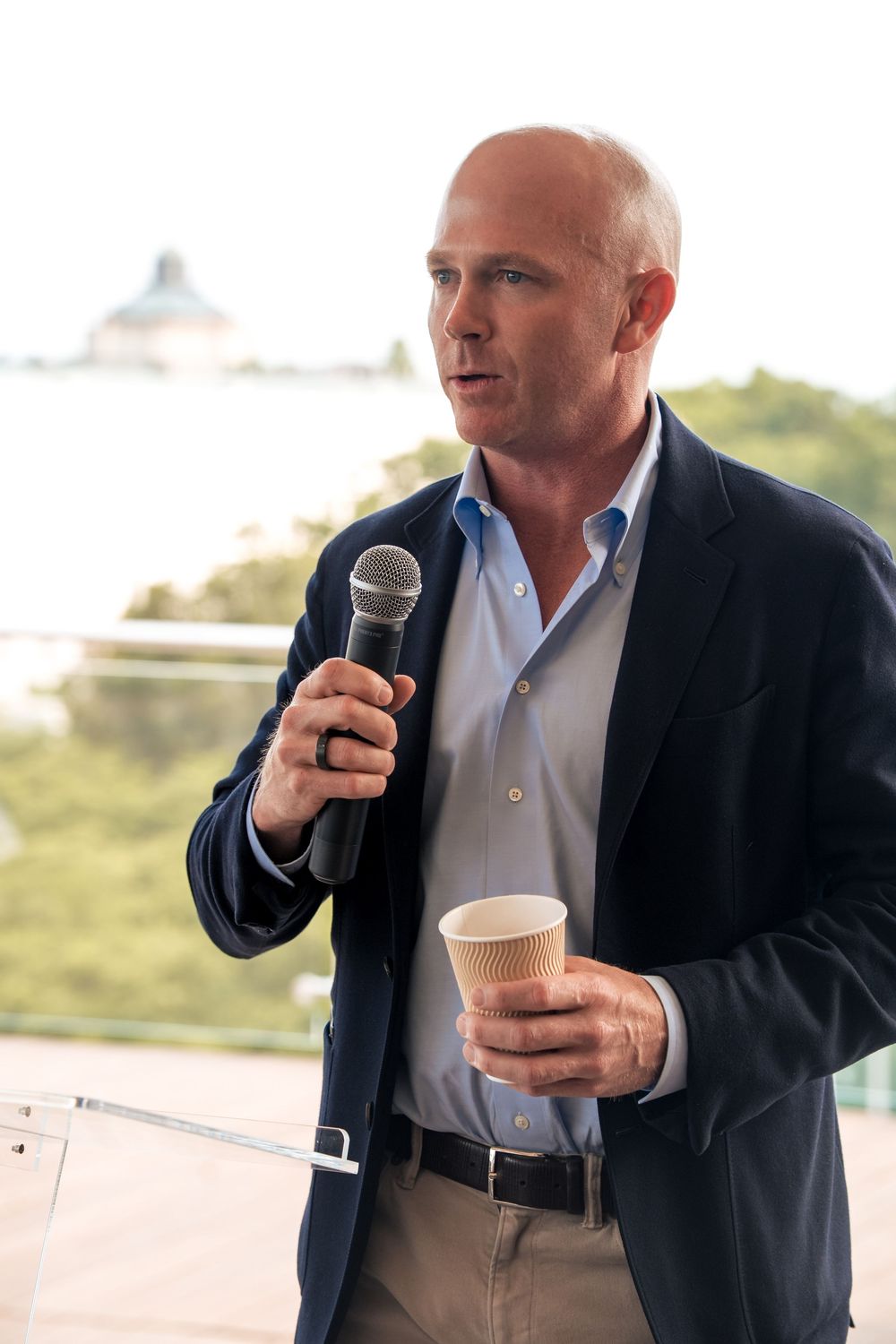 Man in business attire speaking into microphone and holding coffee cup on outdoor terrace.