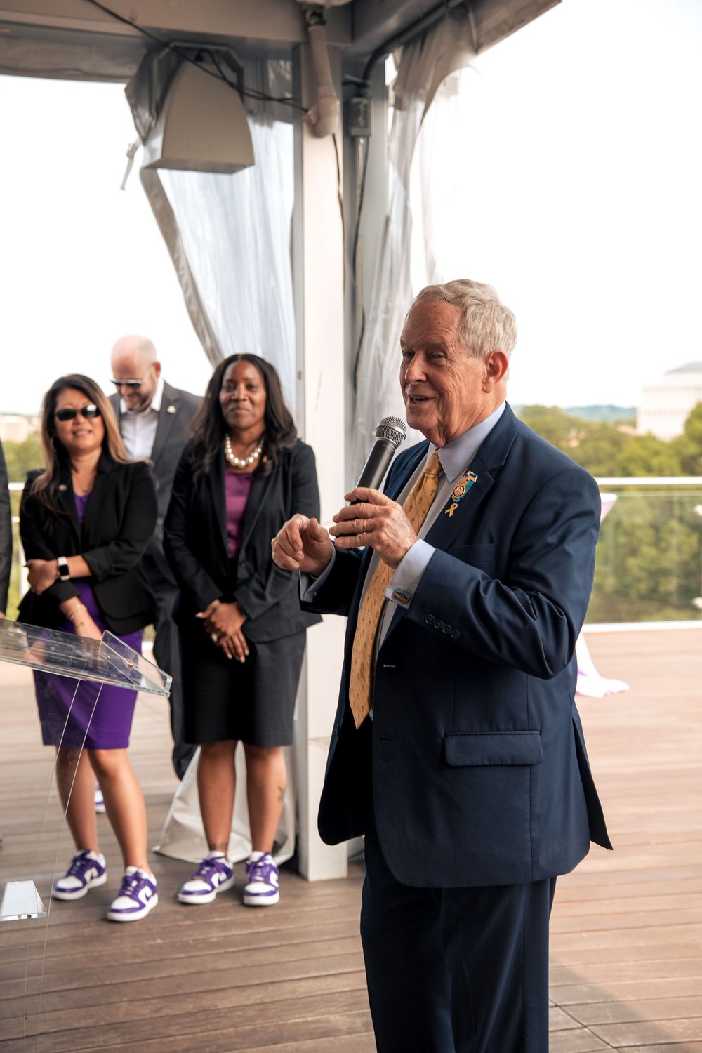 Speaker holding microphone addresses audience on covered outdoor deck with scenic backdrop.