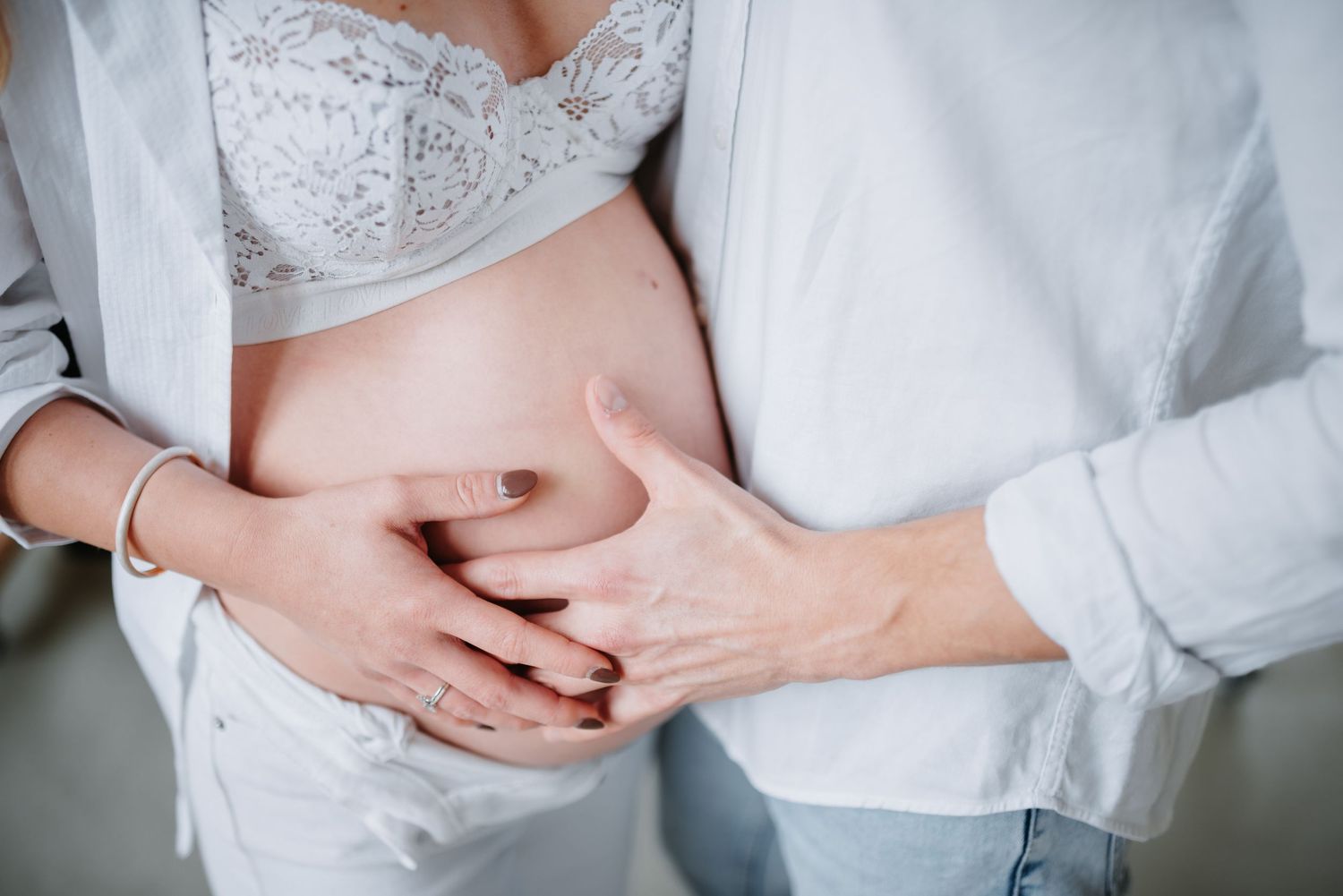 Close-up of hands gently cradling a pregnant belly while wearing a white lace top and gray cardigan.