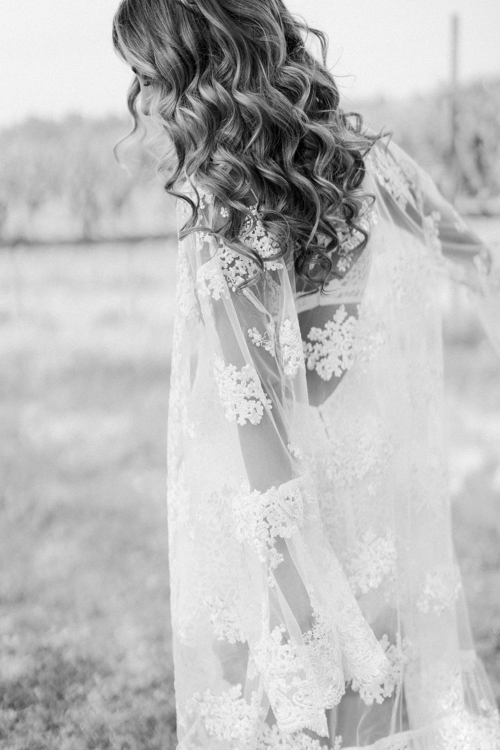 Black and white portrait showing delicate lace wedding veil and flowing curled hair in an outdoor setting.