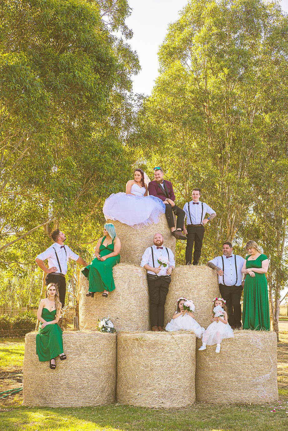 A wedding party poses creatively arranged on stacked hay bales with green emerald dresses and suspenders in an outdoor setting.
