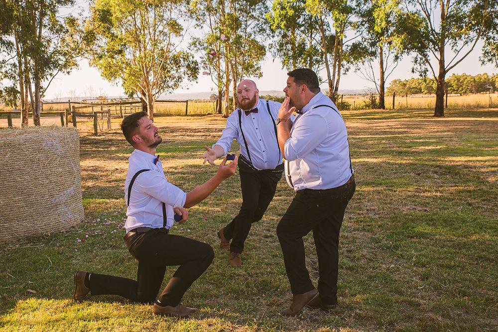 Three people in formal attire dance and laugh together in a sunlit park setting with eucalyptus trees in the background.