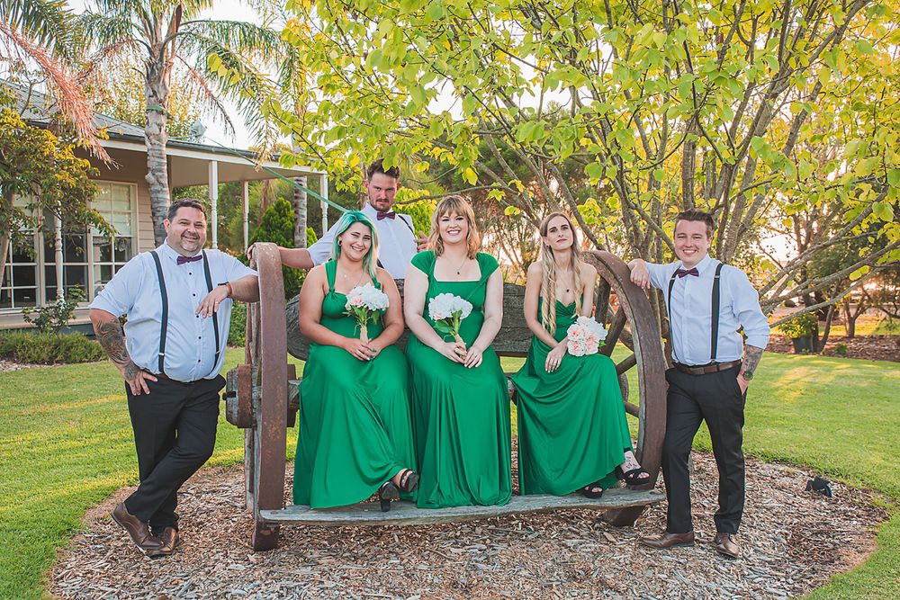 Wedding party in green dresses and suspenders pose playfully together under trees during outdoor photo session.