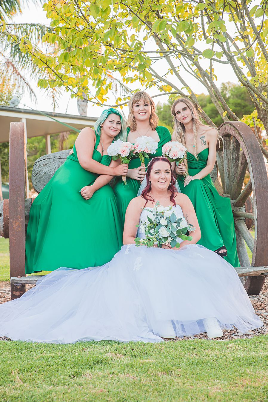 A bridal party in emerald green dresses poses playfully on the grass with the bride in a white gown at sunset.