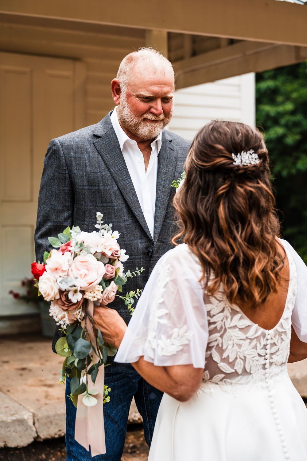 Wedding couple sharing an intimate moment in white dress and grey suit against rustic backdrop.