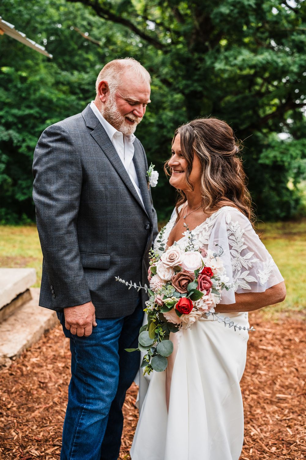 Wedding couple pose outdoors on autumn day, bride holding red and white bouquet with lace dress and groom in grey blazer.