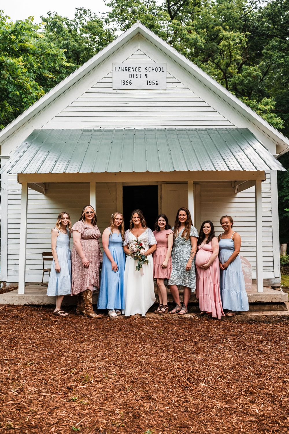 A group in pastel dresses poses in front of a white historic schoolhouse building.