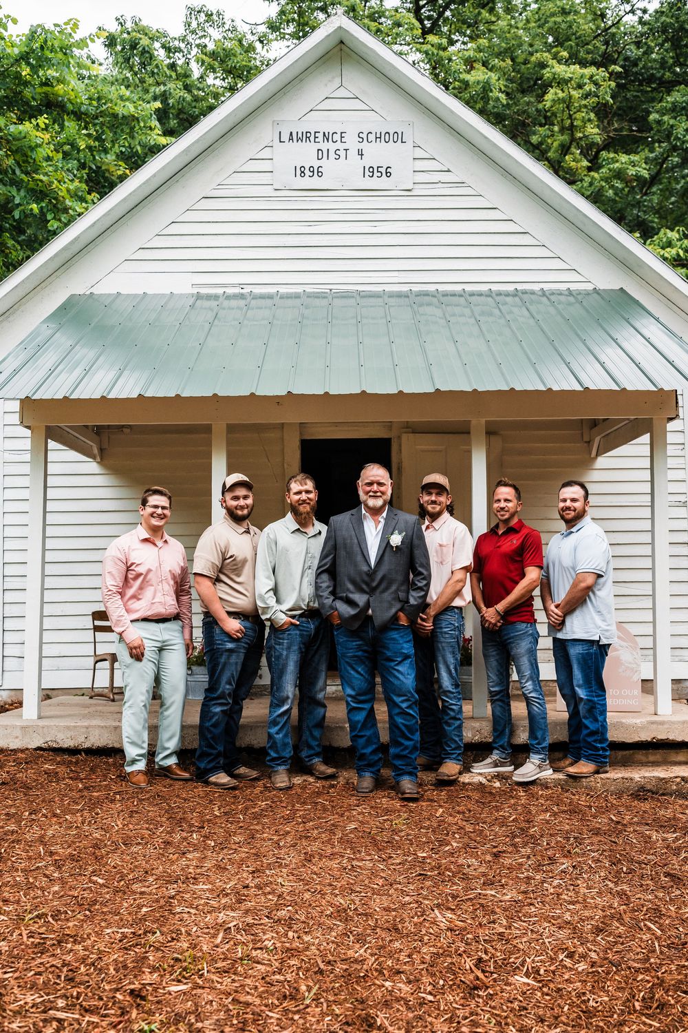 Wedding party members in casual attire lined up against white schoolhouse exterior.