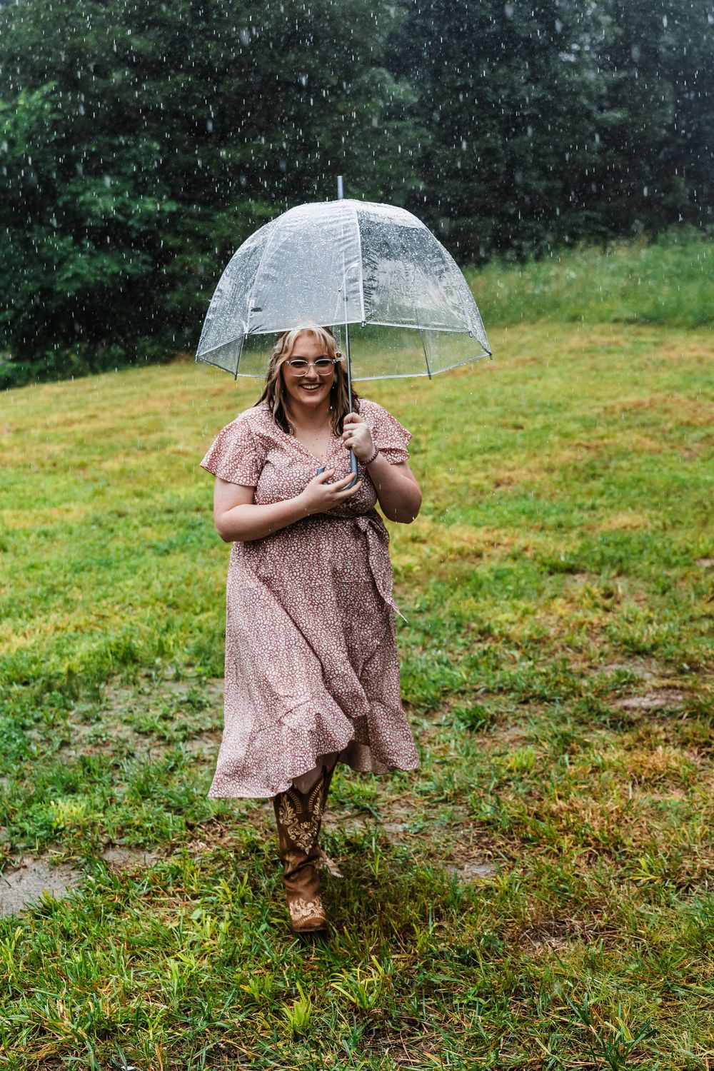Someone in a polka dot dress dances joyfully with a clear umbrella in the rain on a grassy field.