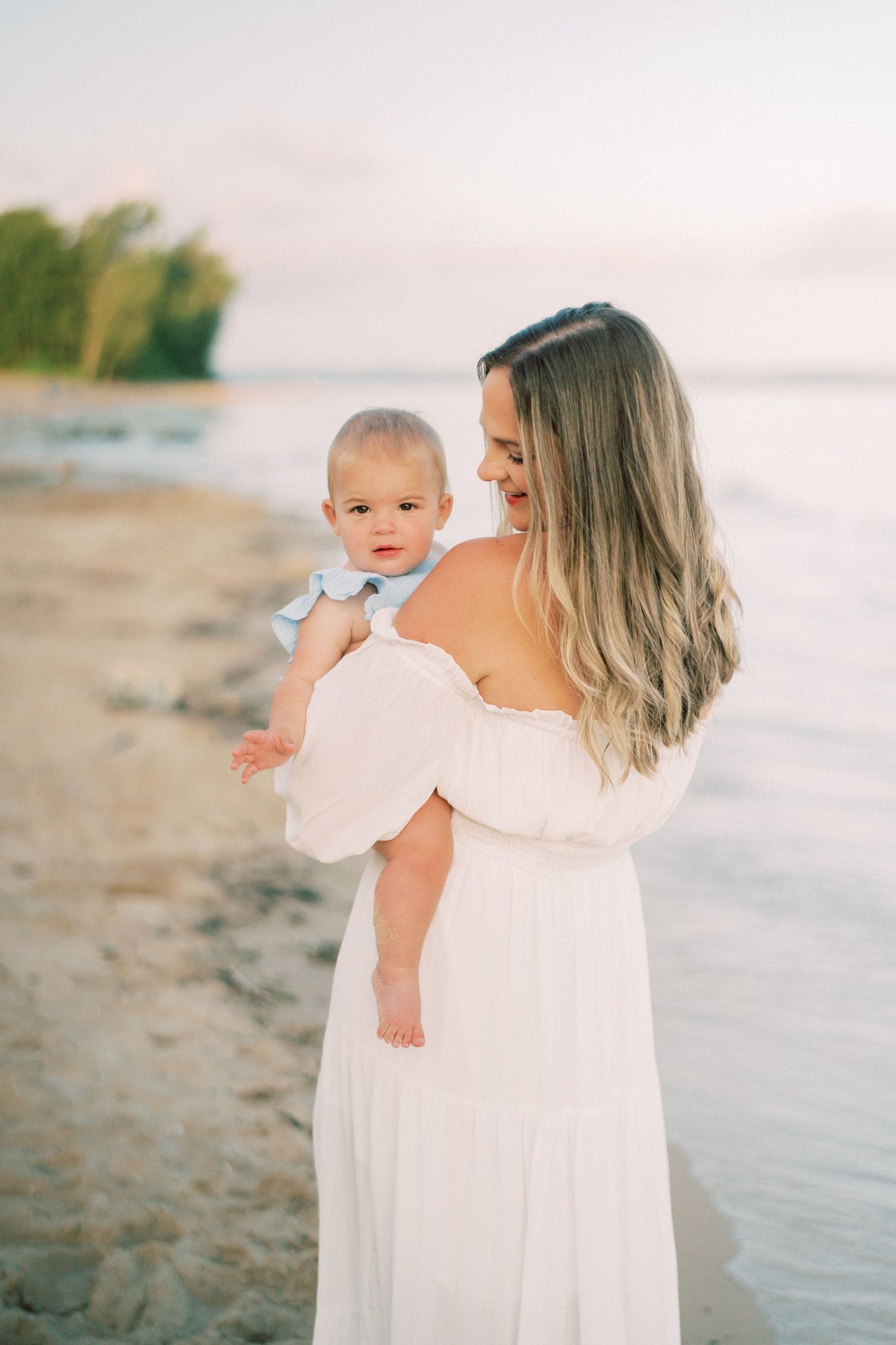 A parent and child share a tender moment together on a peaceful beach at sunset in matching white outfits.