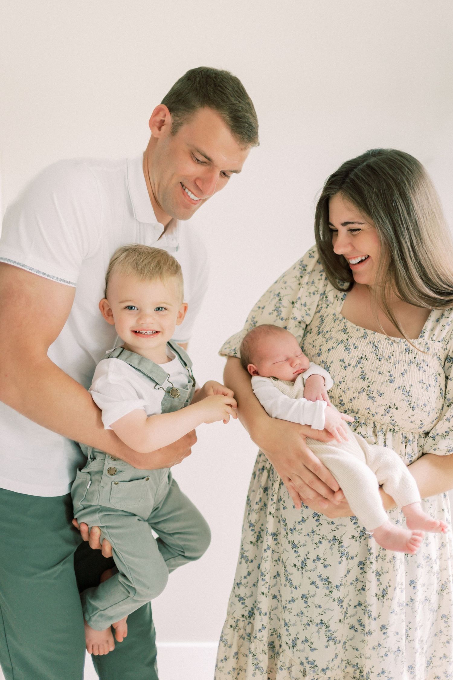 Family of four with floral dress and white shirt standing against white wall during indoor portrait session.
