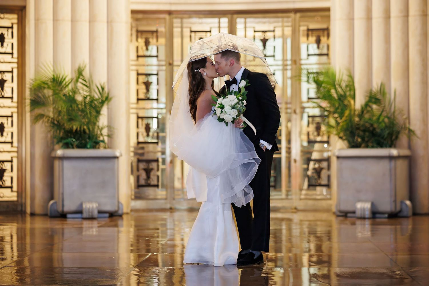 Newlywed couple embraces under an umbrella at historic venue on a rainy day with elegant columns and archways in background.
