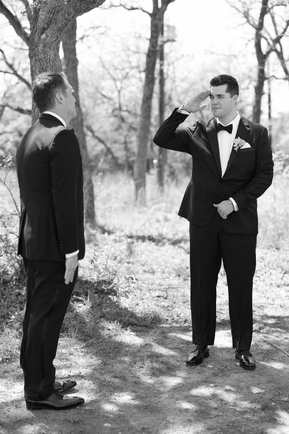 Two men in black tuxedos having a playful moment on a wooded path in black and white photography.