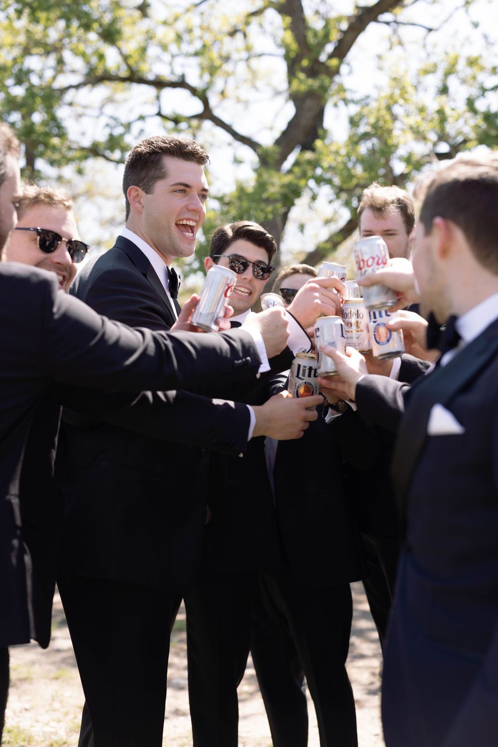 Groomsmen in black suits toasting with beer bottles outdoors during a wedding celebration.