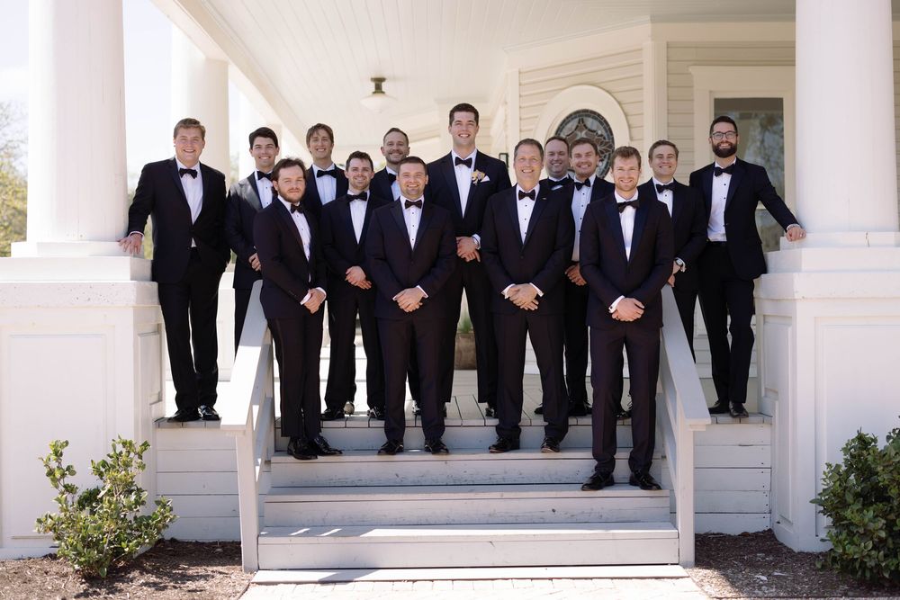 A group of groomsmen in black tuxedos and bow ties stand on white steps outside a traditional columned building.