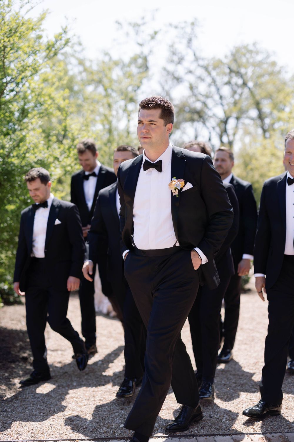 Groomsmen in classic black tuxedos and bow ties walk together outdoors on a sunny day during a wedding event.