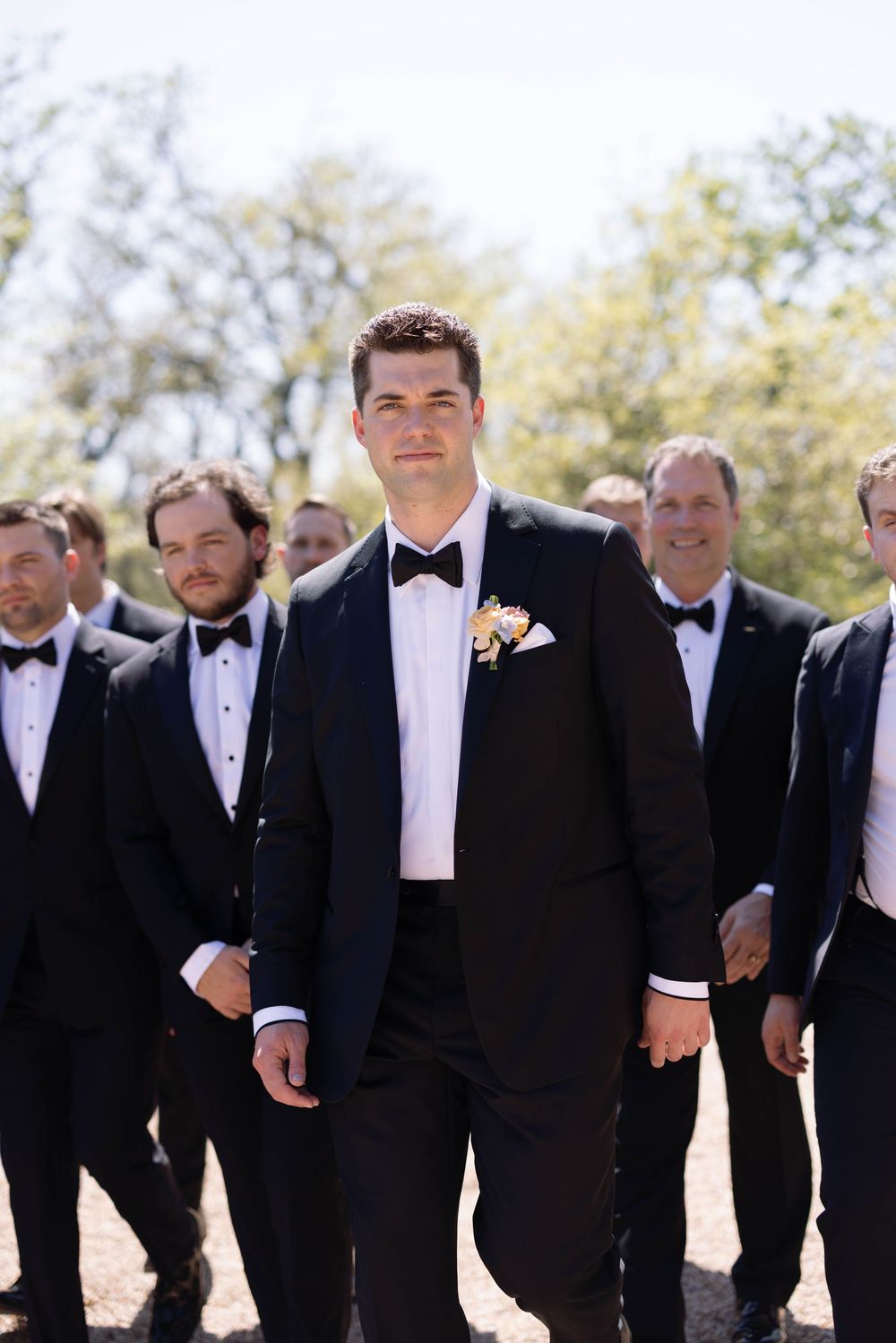 A group of groomsmen in matching black tuxedos and bowties line up outdoors under sunny skies.