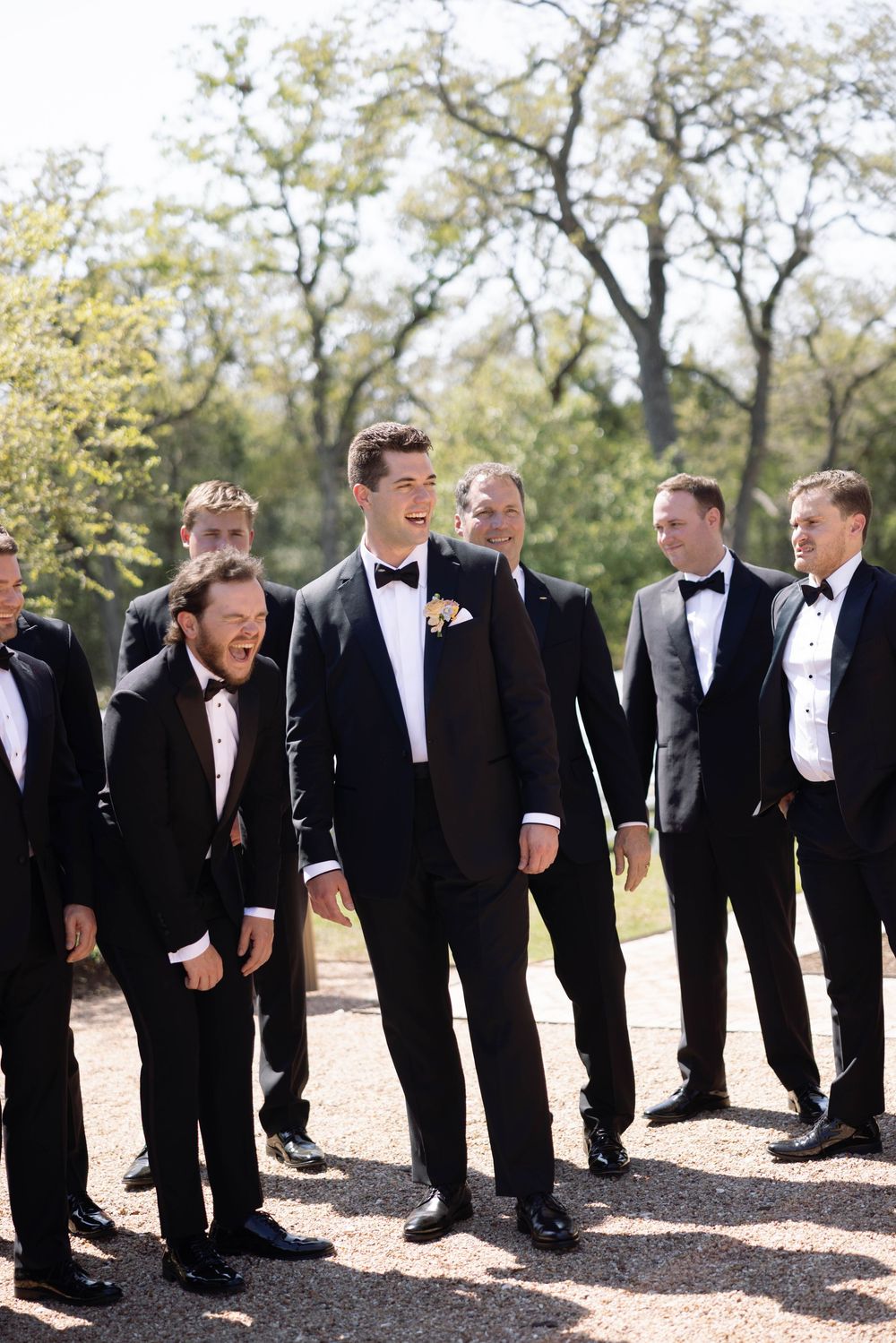A line of groomsmen in black tuxedos with white shirts stand outdoors in bright sunlight, laughing together.