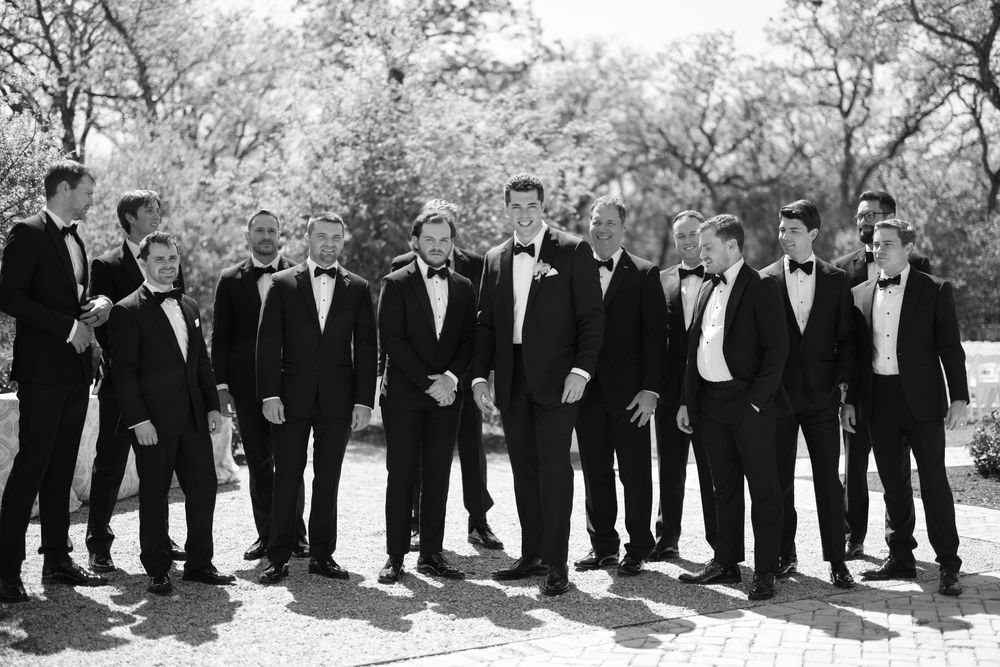 A group of groomsmen in formal black tuxedos standing together outdoors in a black and white photograph.