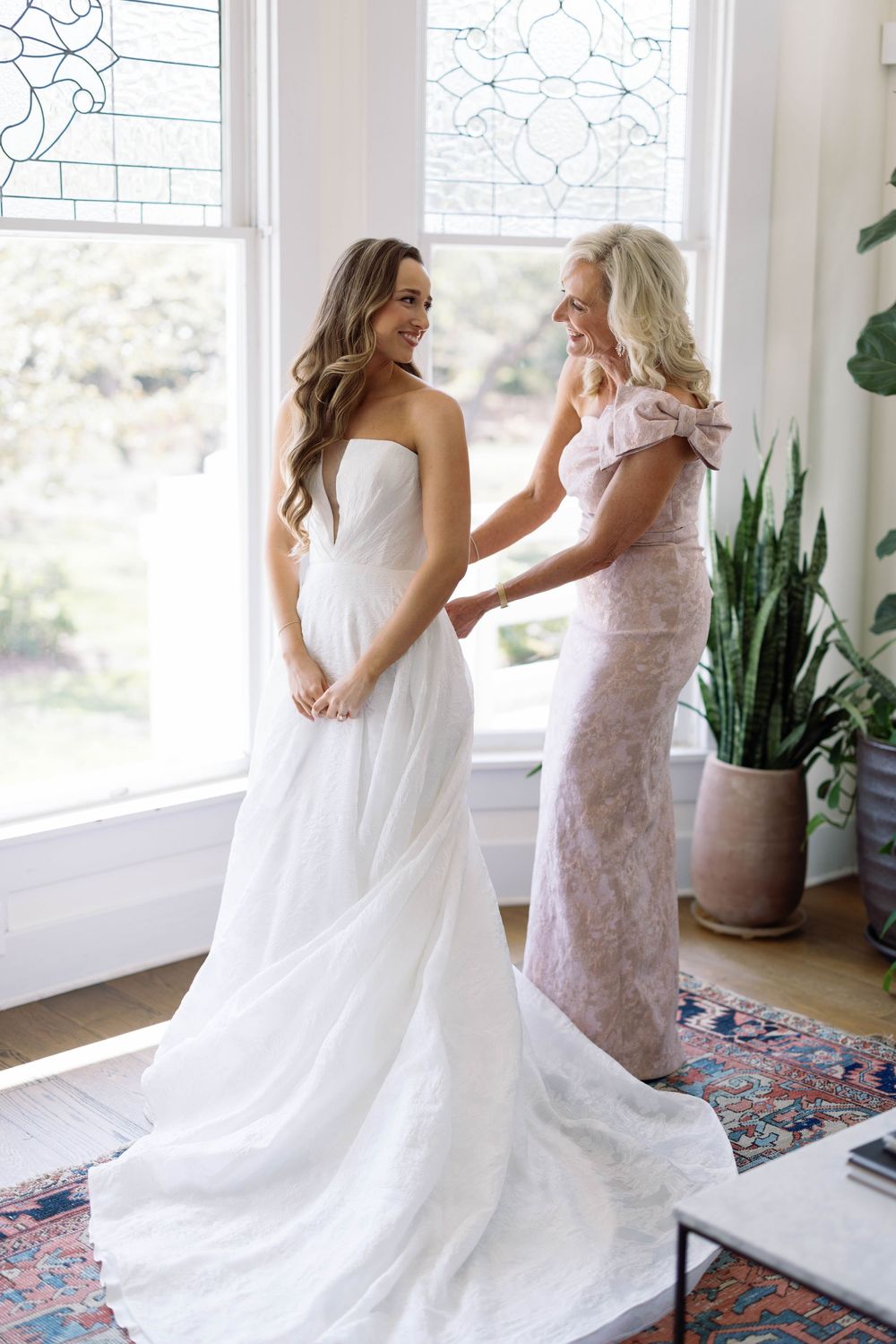 Two people in white and light pink dresses stand together near large windows in a bright room with a patterned rug.