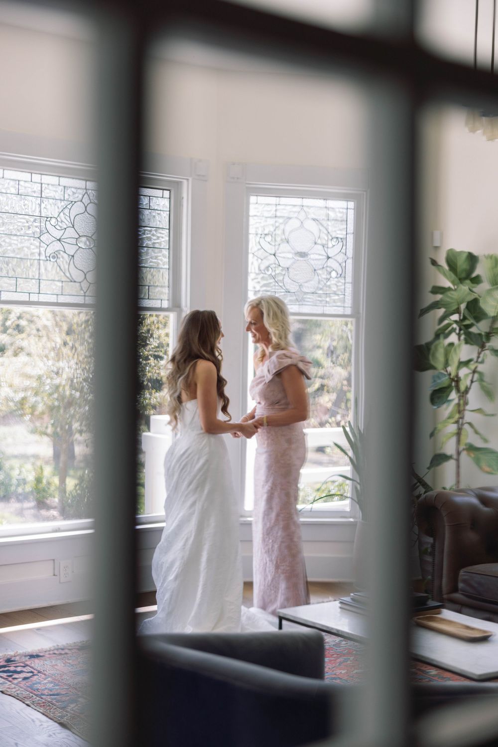 Through window bars, two people in white and pink dresses share an emotional moment in a bright sunlit room.