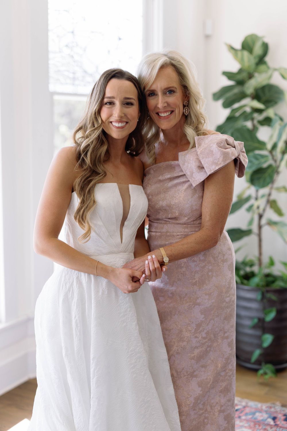 Two people in formal attire share joyful embraces and laughter in a sunlit room with houseplants.