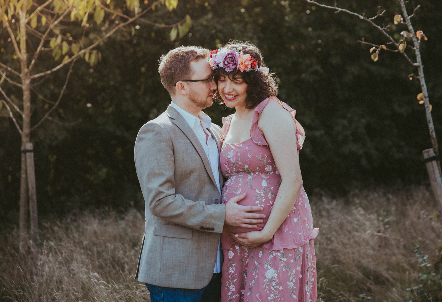 A romantic outdoor maternity photoshoot shows a couple embracing in a field at sunset, with the pregnant woman wearing a pink dress.