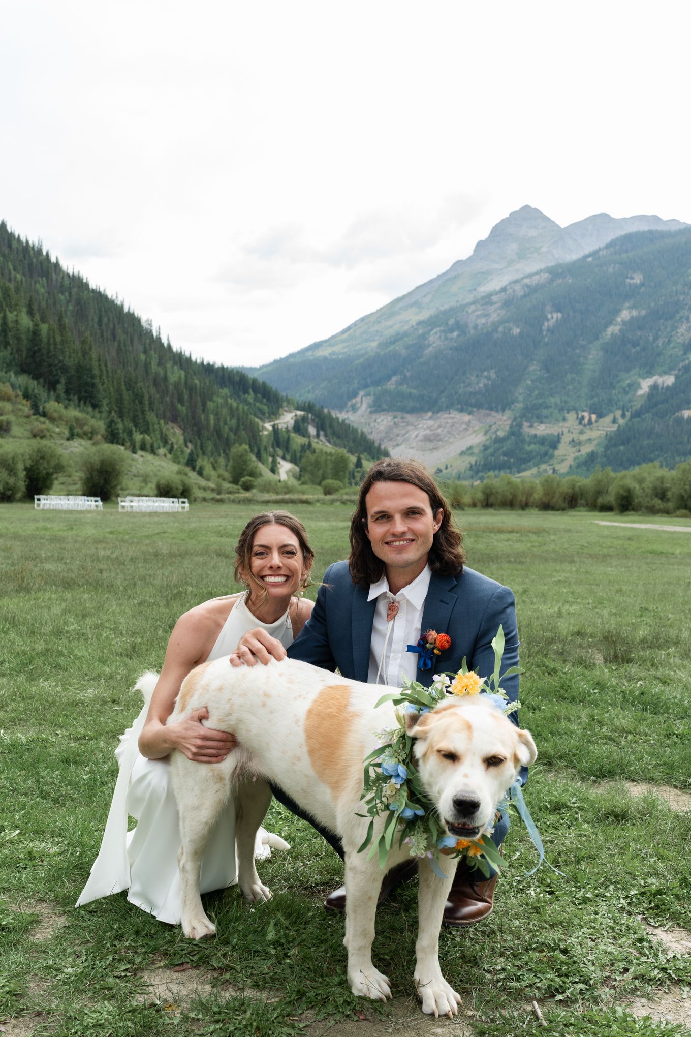 Couple poses with their dog wearing a floral garland in a mountain meadow.