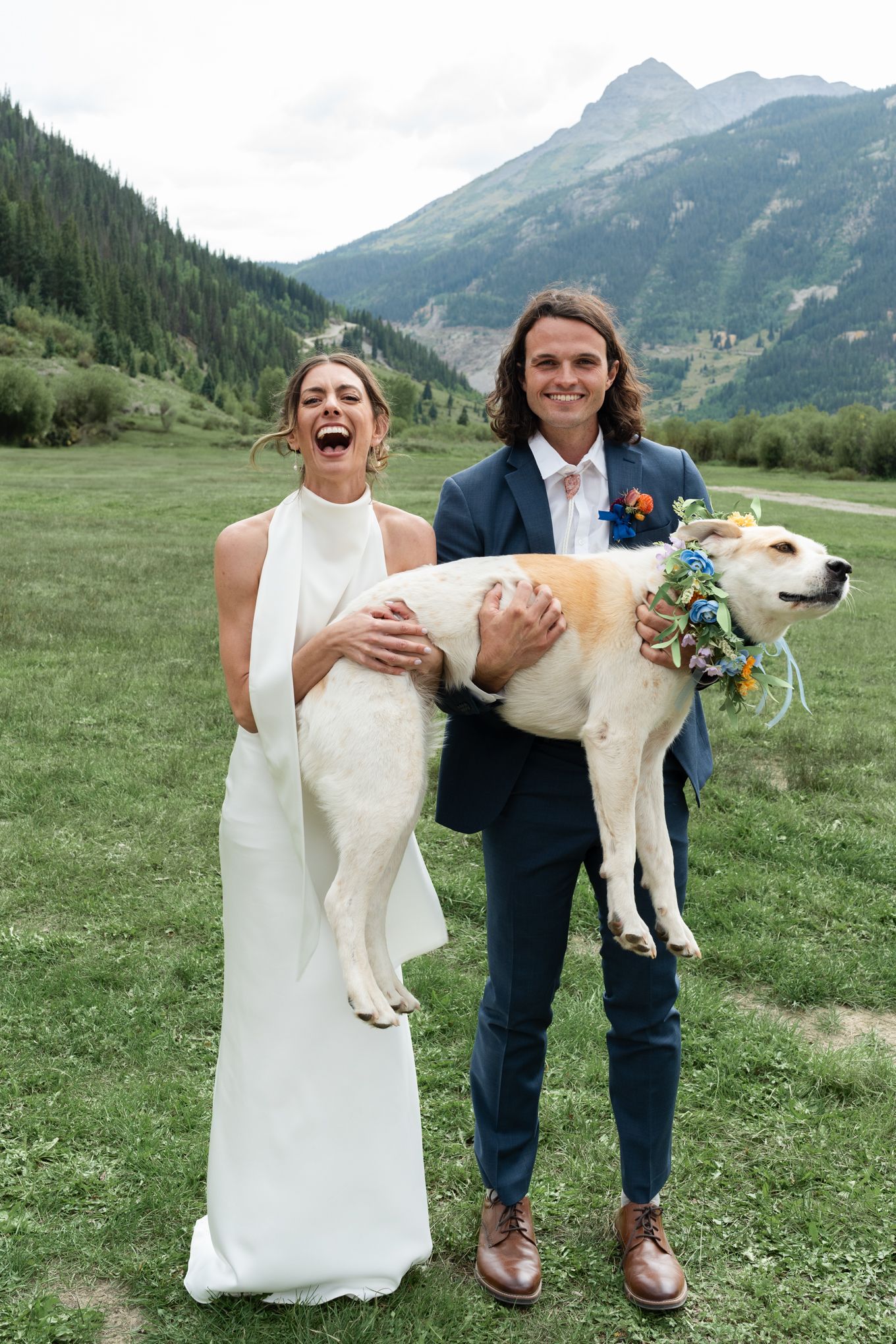 Happy couple lifts their dog while posing in mountain valley setting.