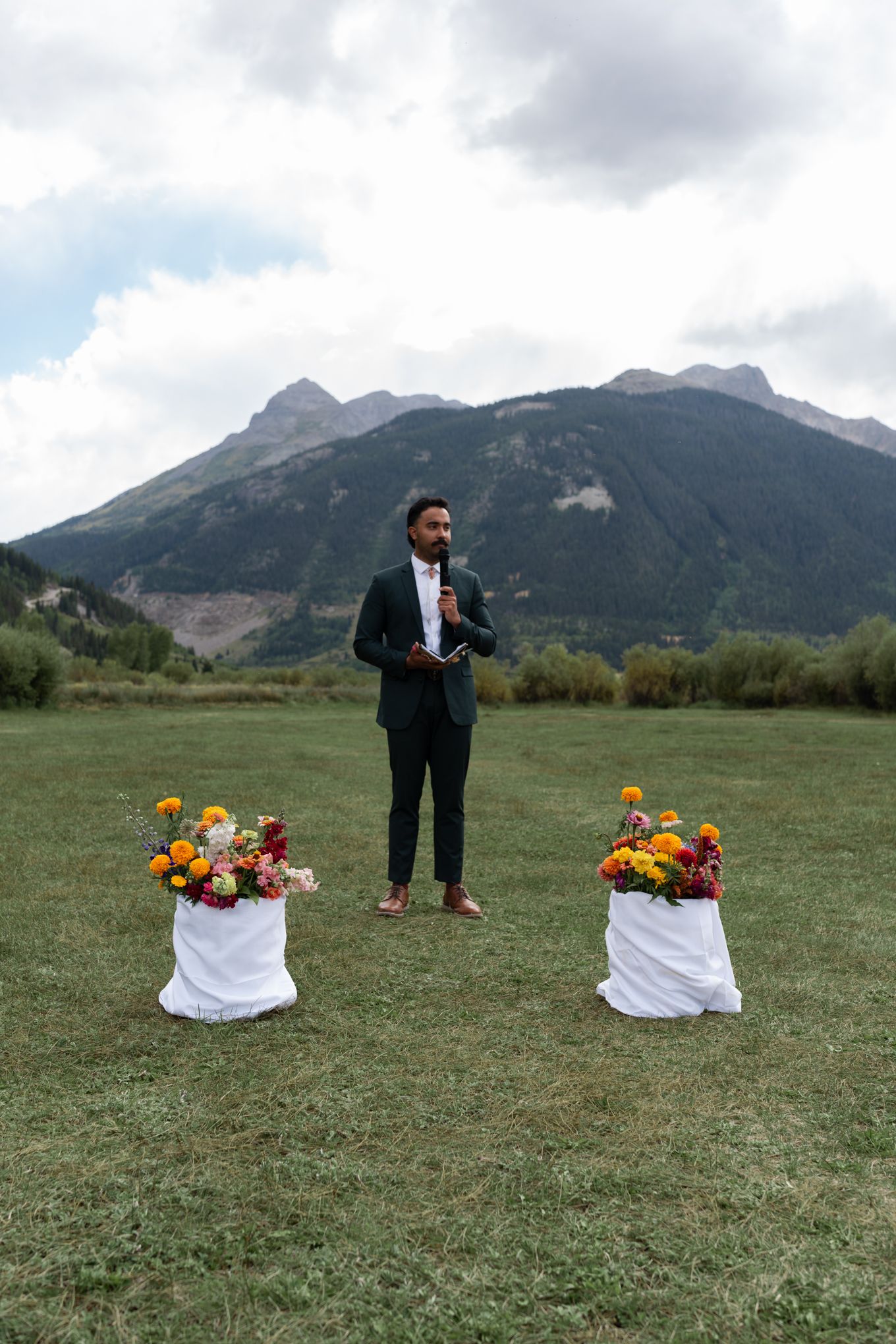 Person in dark suit stands between two floral arrangements with mountain backdrop.