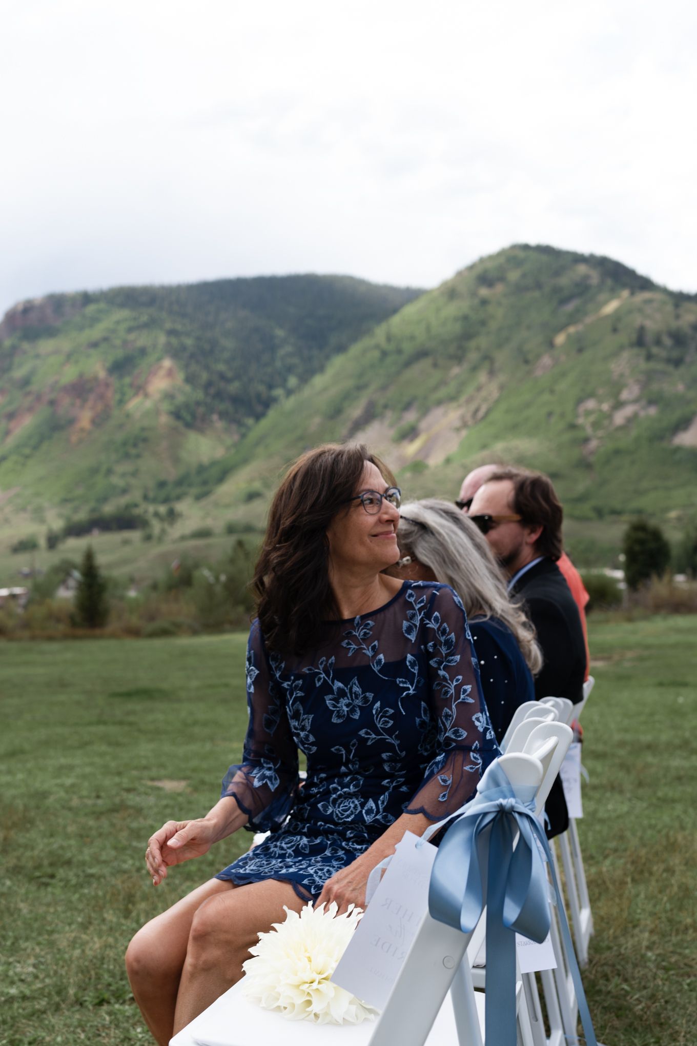 Wedding guest in blue dress sits on white chair at outdoor mountain ceremony.
