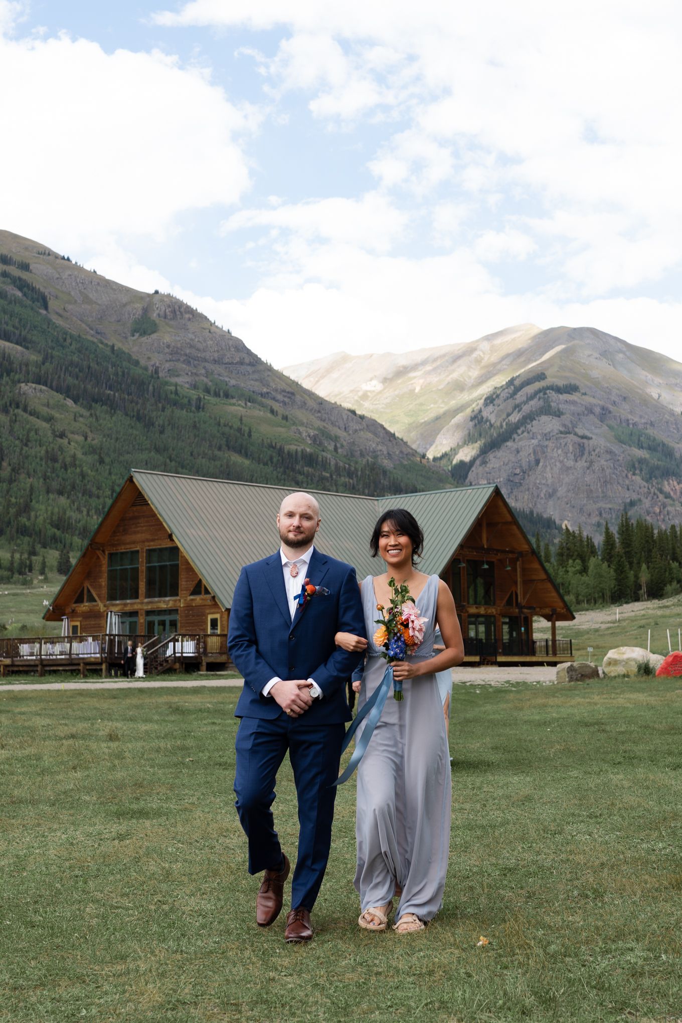 Couple walks on grassy lawn with wooden lodges and majestic mountain peaks in the background.
