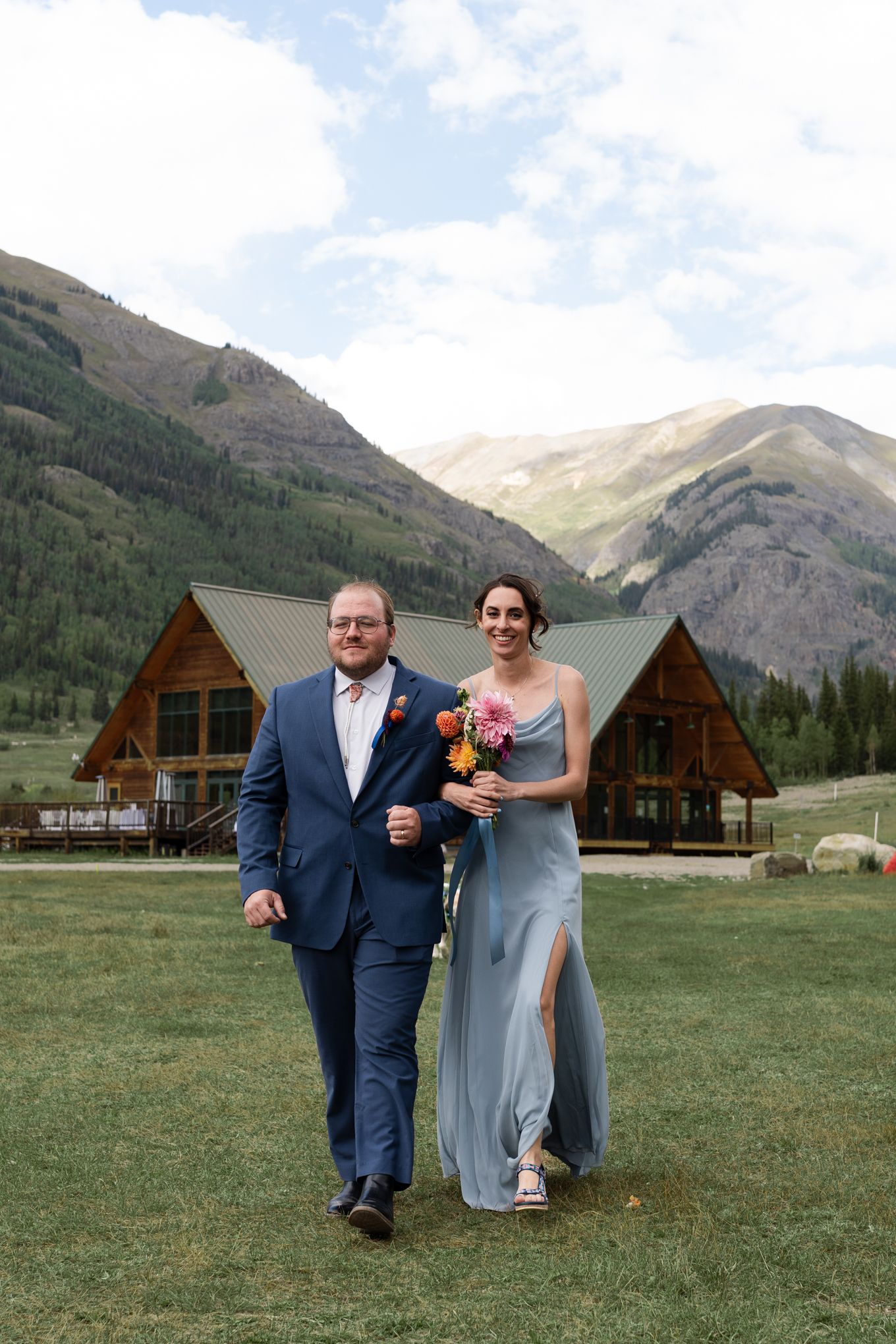 Wedding ceremony participants walk across mountain meadow with rustic log cabins in background.