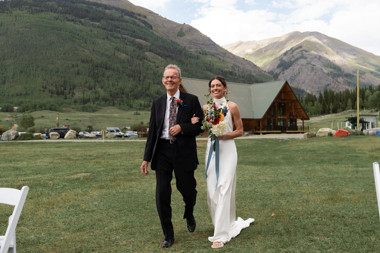 Wedding ceremony on mountain meadow with rustic wooden lodge and peaks in background.