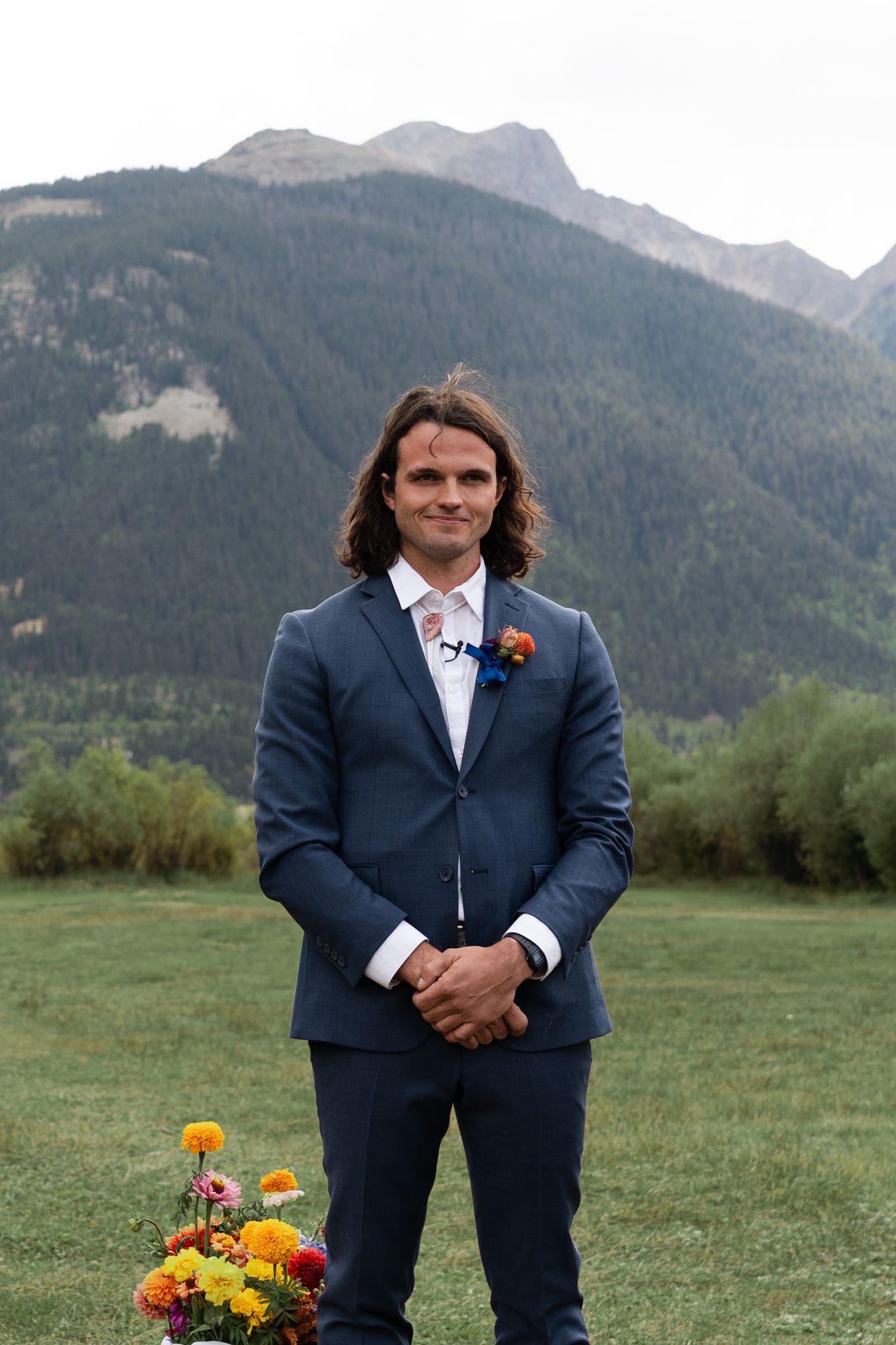 Wedding ceremony participant stands in navy suit with mountain landscape behind.