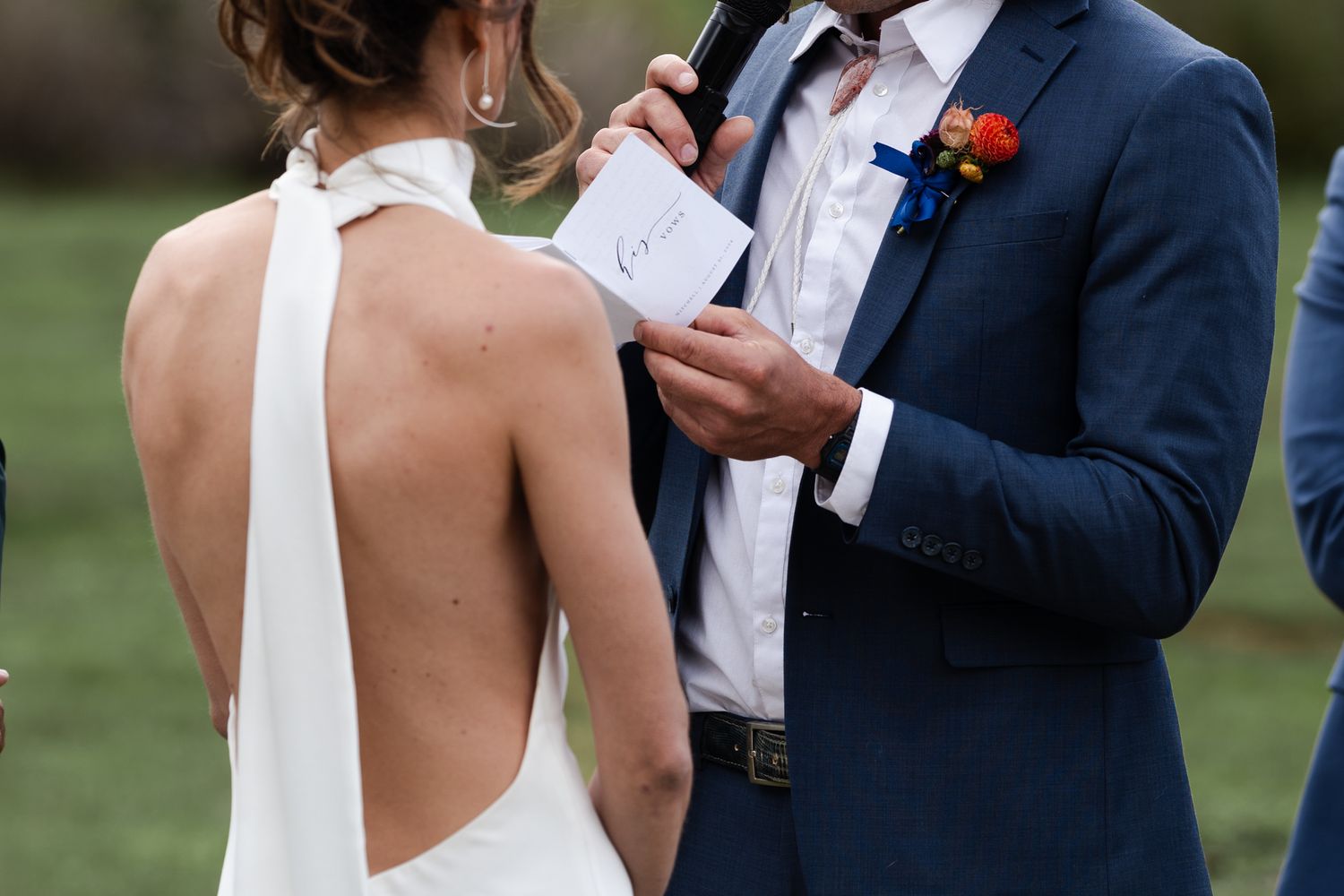Close-up shot of a bride's backless white dress and groom reading vows during outdoor mountain ceremony.