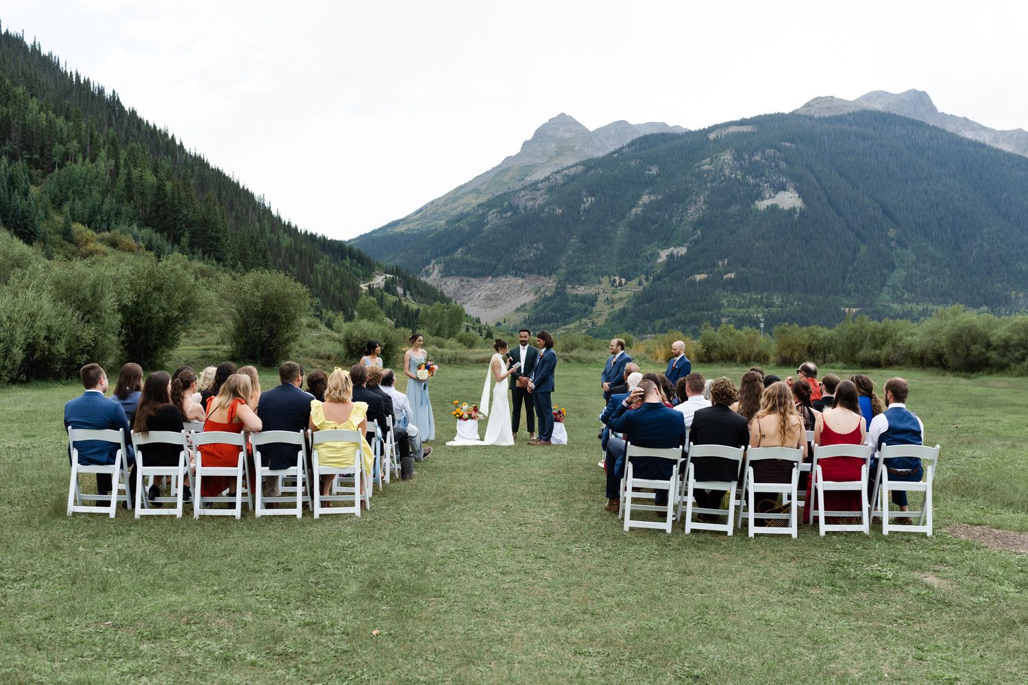 Intimate outdoor mountain wedding ceremony with guests seated in white chairs against dramatic mountain backdrop.