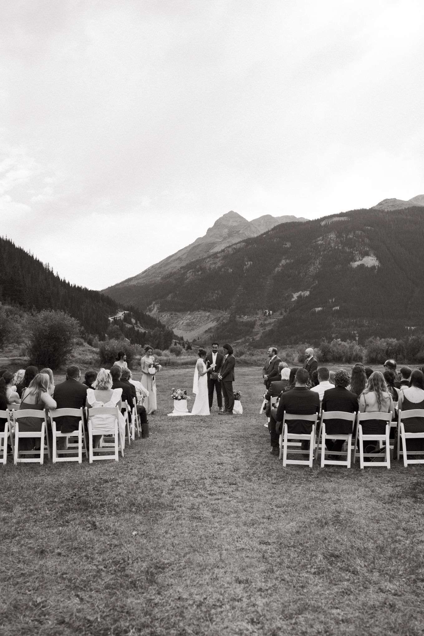 Black and white photo of mountain wedding ceremony with guests seated in two sections on white chairs.