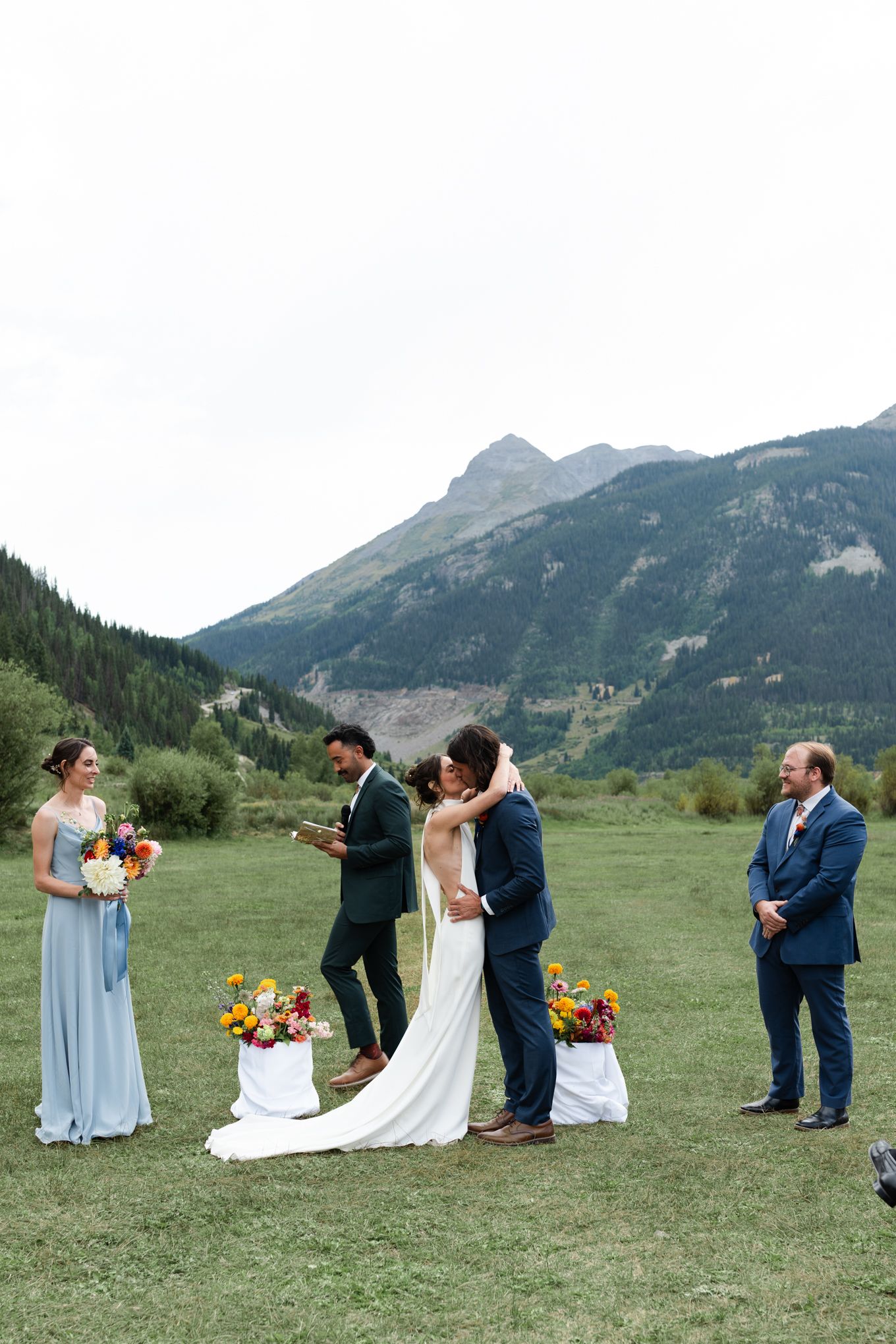 Small wedding party gathered for ceremony with mountain backdrop and colorful floral arrangements.