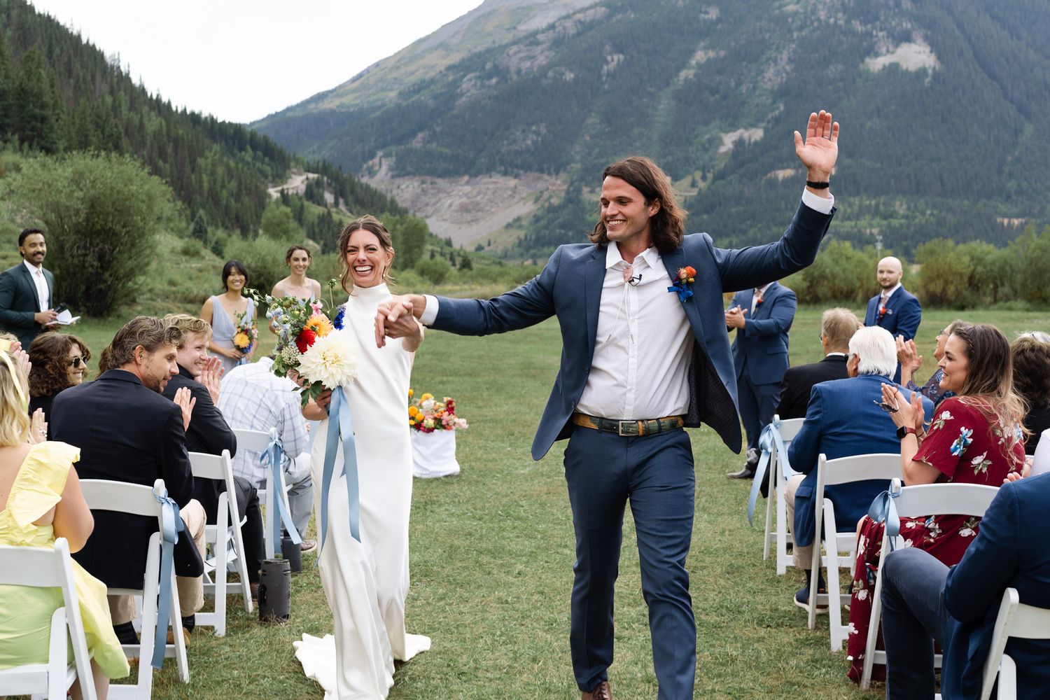 Newlyweds celebrating with raised arms while walking down aisle at mountain wedding ceremony.