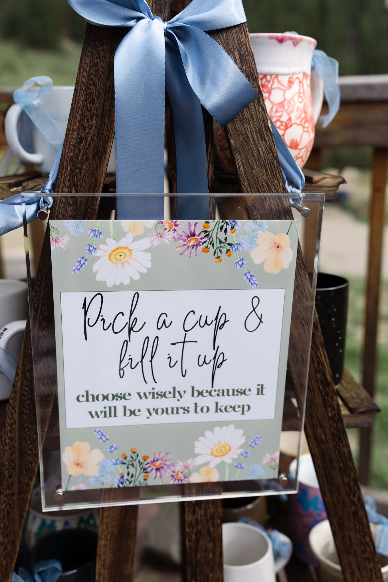 Floral wedding sign reads 'Pick a cup & fill it up' with light blue ribbons on a wooden display.