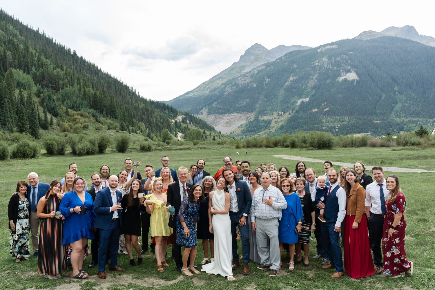 Wedding guests and couple pose together for a panoramic group shot in a mountain valley setting.