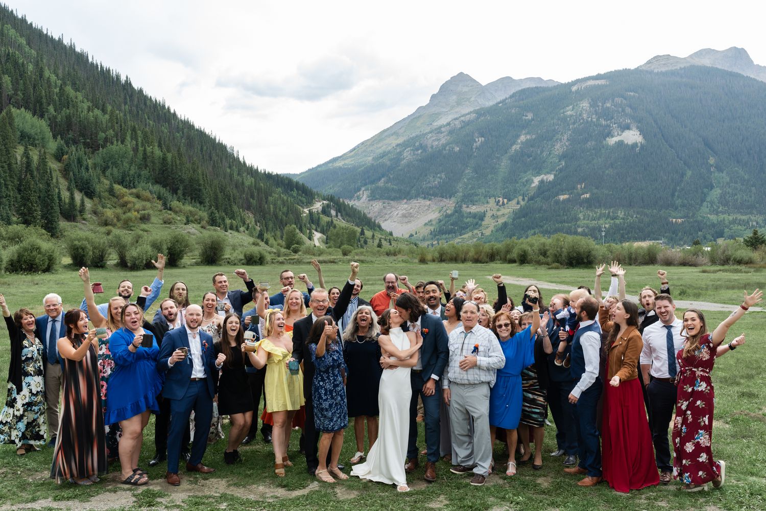 Large wedding party gathered for a group photo with majestic mountains in the background.