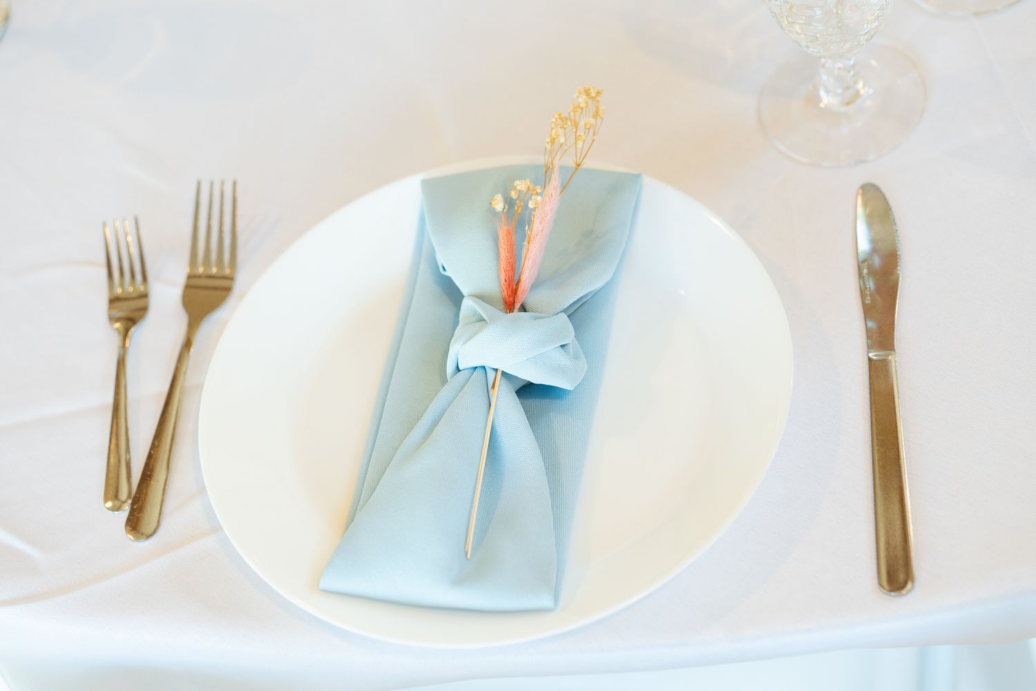 Elegant place setting with light blue napkin fold and gold cutlery on white plate.